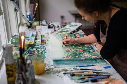 A woman wearing an apron and beaded earrings works at a sunlight desk, painting a picture of children in a field of blue and red berries. Around her are more paintbrushes, containers of paint-colored water and supplies, and palettes of colors.