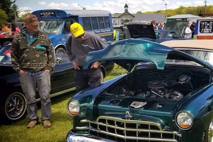 Two men inspect under the hood of a dark teal classic car, parked on a green lawn with other old vehicles.