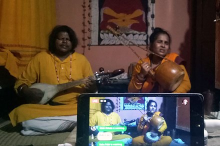 A man and woman perform music sitting on the ground, both holding stringed instruments with their eyes closed. A smartphone in the foreground is live streaming their performance.