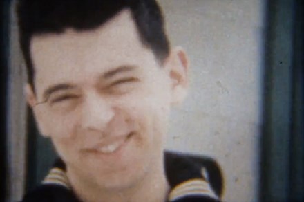 Old film still of a young man with a dark crew cut, in a Navy uniform, smirking at the camera.