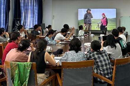 In a classroom, students sit around a large oval table, watching a video screen showing two people outdoors, a microphone set on a tripod between them. 