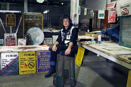 A person wearing black clothes, with glasses and spiked dark hair, sits on an overturned wastebasket in a workshop, surrounded by 'no littering' signs, screenprinting equipment, and prints strung up to dry.