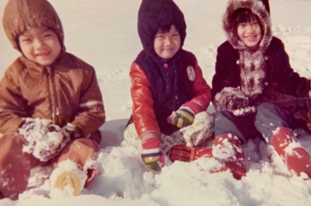 Three small Vietnamese children in hooded jackets, gloves, and boots sit in the snow, smiling.