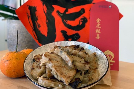 Bowl of pale brown rectangular vegetable cakes, set in front of orange citrus, a red envelope with Chinese characters in gold, and a red paper with a black, hand-painted Chinese character.