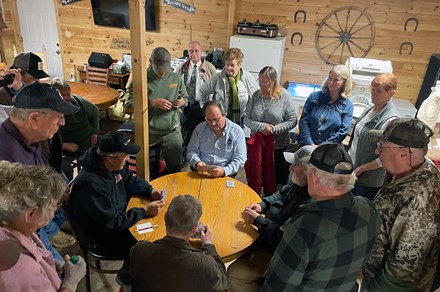 Four men sit around a round wooden table playing cards, while a crowd of others surround them.