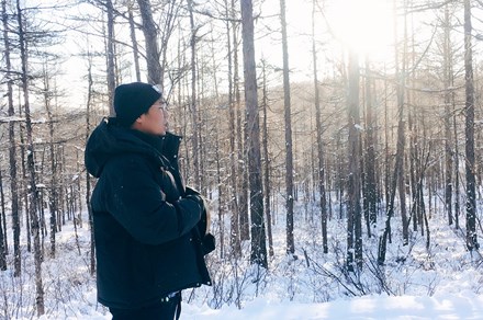 A man in heavy black jacket and beanie stands in front of a bare forest, snow covering the ground, calling out.