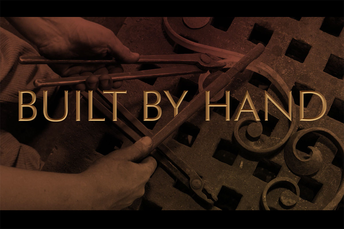 In sepia tone, close-up on a pair of hands using a tool to shape a decorative piece of wrought iron. Overlaid on the image, the words Built by Hand, in a font mimicking a hand-carved inscription.
