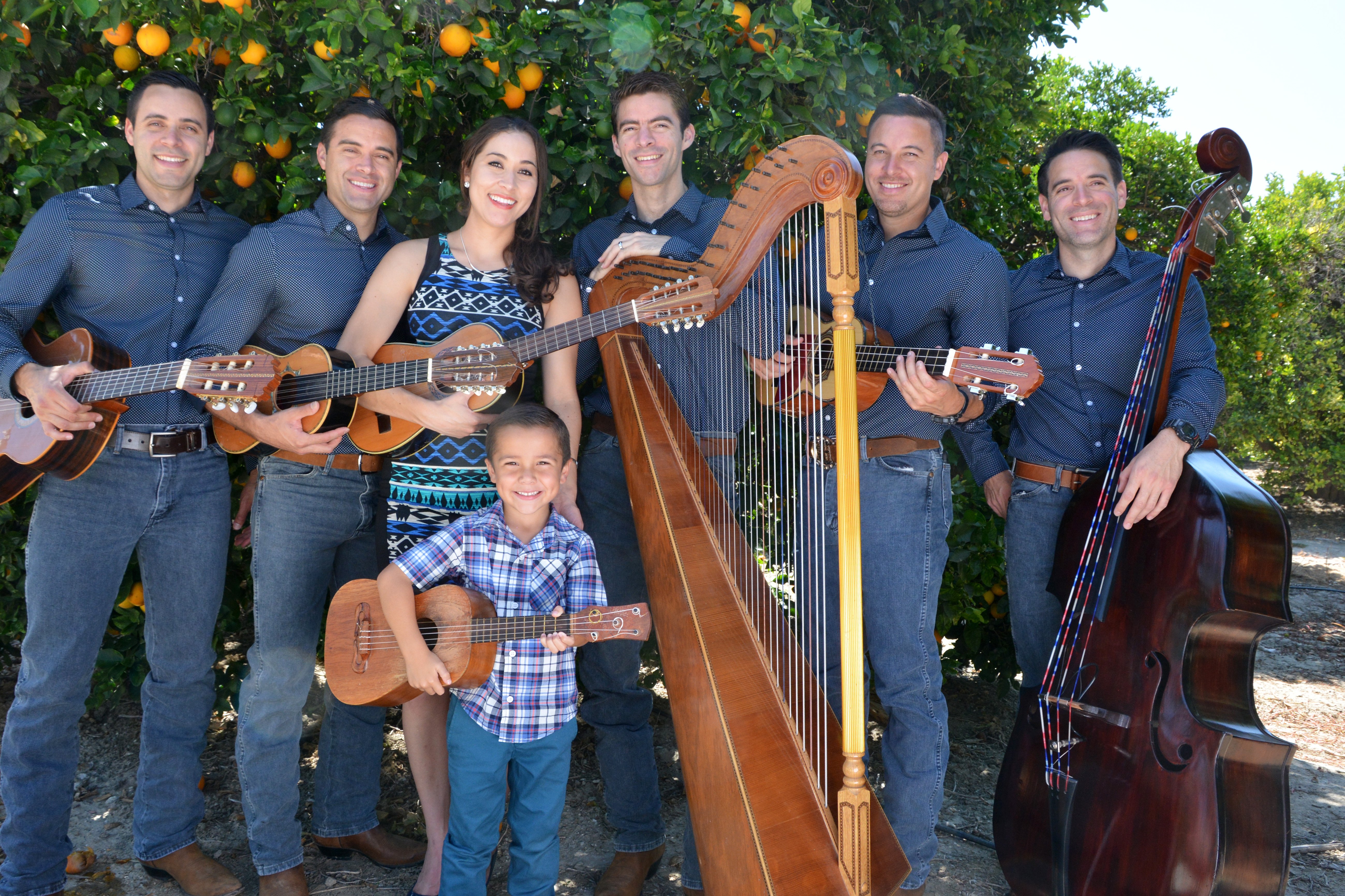 Six adults and a child post with wooden musical instruments in an orange orchard. Instruments include various sizes of guitars, a harp, and an upright bass.